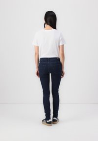 Jeans ajustados de color azul marino fabricados en denim, combinados con una camiseta de algodón blanca. Modelo vistiendo zapatillas negras y blancas, visto desde atrás.