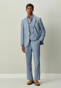 Traje de lino azul claro con blazer y pantalones a juego, con tres botones y una camisa de estampado floral debajo. Mocasines marrones.