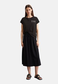 Schwarzes Baumwoll-T-Shirt mit weißem Text "ES IST ZEIT ZU ÄNDERN", kombiniert mit einem langen schwarzen Rock und schwarzen Sandalen, die einen Kordelzugbund haben.