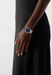 Montre en métal argenté avec un cadran bleu rond, bracelet en acier inoxydable et marqueurs minimalistes, portée sur une main contre un vêtement noir.
