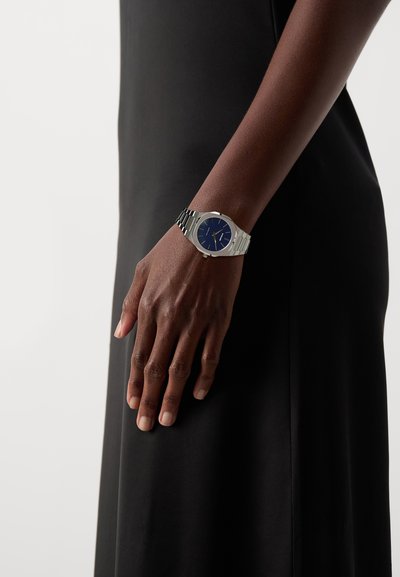 Strieborné kovové hodinky s okrúhlym modrým ciferníkom, oceľovým remienkom a minimalistickými značkami, oblečené na ruke proti čiernemu odevu.