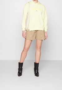 Žlutá mikina s logem "Max Mara", béžové šortky a černé kotníkové boty s detailem přezky. Jednoduchý, ležérní outfit ukazující vrstvené textury.