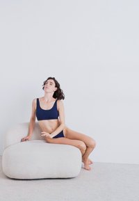 Laivastonsininen crop top ja siihen sopivat bikinit. Malli istuu pehmeällä, pyöreällä, valkoisella teksturoidulla tuolilla yksinkertaista taustaa vasten.