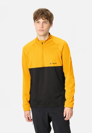 Junger Mann trägt eine gelb-schwarze Langarm-Quarter-Zip-Jacke und schwarze Hose, steht vor einem schlichten weißen Hintergrund.