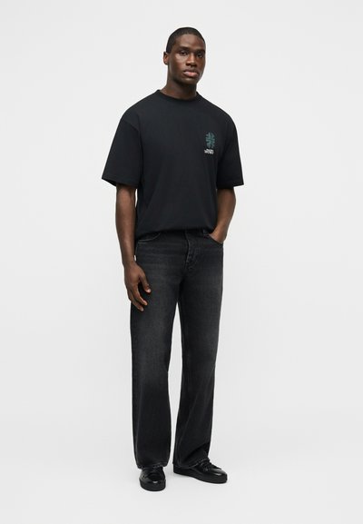 Άνδρας που στέκεται με το ένα χέρι στην τσέπη, φοράει μαύρο μπλουζάκι με σχέδιο, σκούρα τζιν φαρδιάς γραμμής και μαύρα παπούτσια σε απλό φωτεινό φόντο.