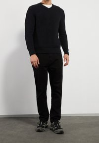 Granatowy sweter z prążkowanego materiału z dekoltem w kształcie litery V, w zestawieniu z czarnymi spodniami i sportowymi butami z zielonymi akcentami. Model stoi na neutralnym tle.