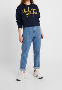 Felpa navy con la scritta "Uniforme dei Liberi" in corsivo giallo, abbinata a jeans in denim azzurro a vita alta con risvolti e sneakers bianche.