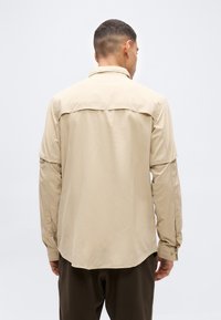 Άνδρας με την πλάτη στραμμένη, φοράει μπεζ πουκάμισο με μακριά μανίκια και κουμπωτά μανσέτα και σκούρο παντελόνι, μπροστά σε ένα απλό φωτεινό φόντο.