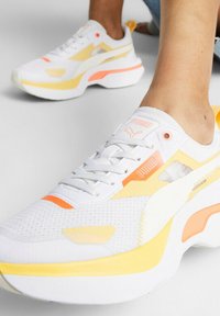Baskets blanches de sport avec des accents jaunes et orange, dessus en mesh, semelle épaisse, design à lacets, et détail de marque proéminent sur la languette.