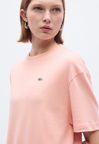 Ροζ ανοιχτό ροζ κοντομάνικο μπλουζάκι από απαλό βαμβάκι με στρογγυλή λαιμόκοψη και μικρό πράσινο κεντητό λογότυπο στο στήθος. Λείο ύφασμα.