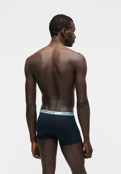 Λεπτός άνδρας με σκουρόχρωμο δέρμα, που στέκεται με την πλάτη στην κάμερα, φορώντας μαύρα boxer briefs Emporio Armani με λευκή μέση, μπροστά σε απλό ανοιχτό φόντο.