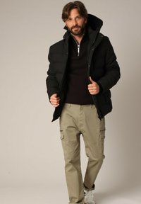 Zwarte pufferjas met een ritssluiting en capuchon, gecombineerd met lichtbeige cargobroek met zijzakken, gedragen met witte sneakers.