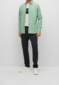 Camicia verde chiaro a maniche lunghe sopra una t-shirt bianca con grafica, abbinata a pantaloni scuri e sneakers bianche. Tessuto morbido, design del colletto classico.