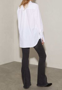 Chemise blanche à col rabattu avec des manches longues, portée déboutonnée, associée à un pantalon évasé gris foncé. Le tissu semble lisse, avec une coupe décontractée.