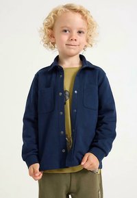 Marineblå skjorte med knapper, to brystlommer, lange ærmer og en glat tekstur, båret over en lysegrøn grafik T-shirt.