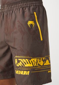 Calções masculinos em tecido castanho-escuro com acabamento brilhante. Apresentam detalhes em amarelo, um bolso lateral com fecho e um logótipo de cobra. Detalhe de padrão abstrato.