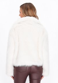 Kvinde vendt væk iført en hvid faux fur jakke og mørkebrun læderbukser mod en hvid baggrund.