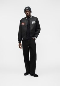 Μαύρο μπουφάν bomber με λευκές ρίγες, κεντημένα μπαλώματα, συνδυασμένο με μαύρο παντελόνι και σκουφάκι. Το μοντέλο στέκεται μπροστά σε ουδέτερο φόντο.