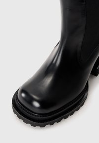 Botte de cheville en cuir noir avec semelle crantée épaisse et panneau élastique sur les côtés, présentée en gros plan en angle sur un fond clair.