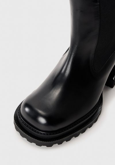 Botte de cheville en cuir noir avec semelle crantée épaisse et panneau élastique sur les côtés, présentée en gros plan en angle sur un fond clair.