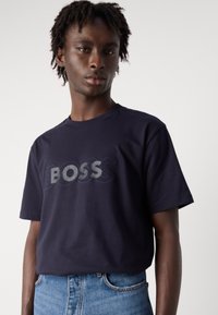 Tamsiai mėlynas medvilnės marškinėliai su ryškiu "BOSS" logotipu įvairiais atspalviais. Trumpomis rankovėmis, apvaliu kaklu, laisvo kirpimo, derinami su šviesia džinsine medžiaga.