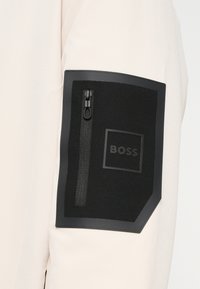 Μανίκι σε ανοιχτό μπεζ χρώμα με μαύρο ταμπελάκι· διαθέτει τσέπη με φερμουάρ και το λογότυπο "BOSS" ανάγλυφο στο ταμπελάκι. Ομαλή υφή υφάσματος.