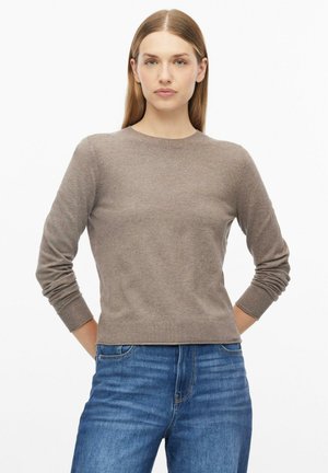 Vrouw met steil lichtbruin haar die een effen bruine trui met lange mouwen en blauwe spijkerbroek draagt, staat met haar handen achter haar rug.