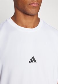 Weißes Sport-T-Shirt aus strukturiertem Stoff, mit einem kleinen schwarzen dreieckigen Logo auf der Brust und Rundhalsausschnitt.