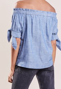 Top azzurro chiaro, scollato sulla spalla, realizzato in un tessuto morbido e texture, con maniche a sbuffo e dettagli di fiocchi, e un collo arricciato.