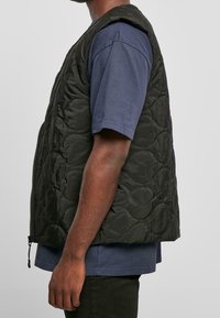 Boční pohled na osobu oblečenou v černé prošívané vestě přes modrou košili s krátkým rukávem a černé kalhoty na jednotném pozadí.