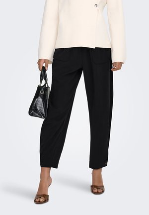 Persona que lleva pantalones negros recortados, top crema con botones, tacones marrones abiertos en la punta y sostiene un pequeño bolso negro.