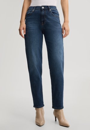Donna con jeans blu scuro a vita alta e gamba dritta e stivaletti beige con punta appuntita, in piedi su uno sfondo semplice.
