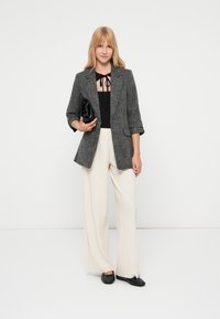 Blazer xadrez preto e branco por cima de uma blusa preta, combinado com calças largas creme e sapatos pretos rasos. A carregar uma carteira preta.