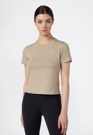 Junge Frau mit dunklen Haaren, trägt ein beigefarbenes Kurzarmoberteil und schwarze enganliegende Leggings, steht vor einem schlichten weißen Hintergrund.