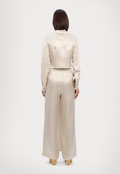 Light beige, satin jumpsuit with long sleeves and wide legs. Features a cropped design and subtle texture. Worn with platform shoes.