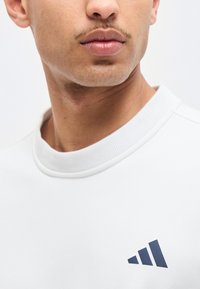 Κοντινό πλάνο του κάτω μέρους του προσώπου και του λαιμού ενός άνδρα που φοράει λευκό μπλουζάκι με στρογγυλή λαιμόκοψη και μικρό μαύρο γεωμετρικό λογότυπο στο στήθος.