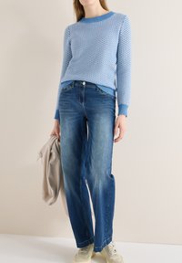Femme portant un pull bleu à motifs, un jean droit de couleur moyenne, une veste beige à la main et des baskets crème, debout contre un mur uni.