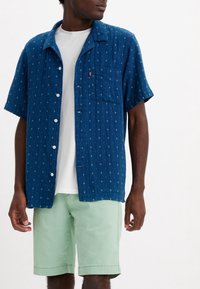 Koszula z krótkim rękawem, zapinana na guziki, w ciemnoniebieskim materiale z małymi białymi wzorami, z kieszonką na piersi i klasycznym kołnierzykiem. W połączeniu z miętowymi szortami.