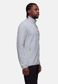 Giacca in pile grigia chiara con zip e colletto alto, due tasche frontali con zip e tessuto testurizzato. Presenta un sottile logo del marchio sul petto.