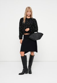 Czarny sweter o textured z kwiatowymi aplikacjami, dopasowana czarna spódnica, wysokie czarne skórzane kozaki i czarna torebka o strukturalnym kształcie.