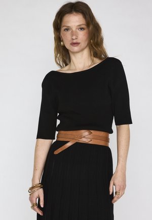 Femme aux cheveux auburn ondulés portant une robe noire côtelée avec des manches au coude et une large ceinture en cuir beige, accessoirisée de bijoux en or.