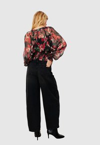 Blouse fleurie noire transparente avec motifs rouges et verts, taille élastique, associée à un jean noir à jambes larges et des bottes noires à talons hauts.