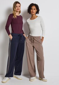 Twee vrouwen in langmouwige tops; de een in bordeauxroo, de ander in wit. Beiden dragen losse sweatpants—marineblauw met witte strepen en beige.