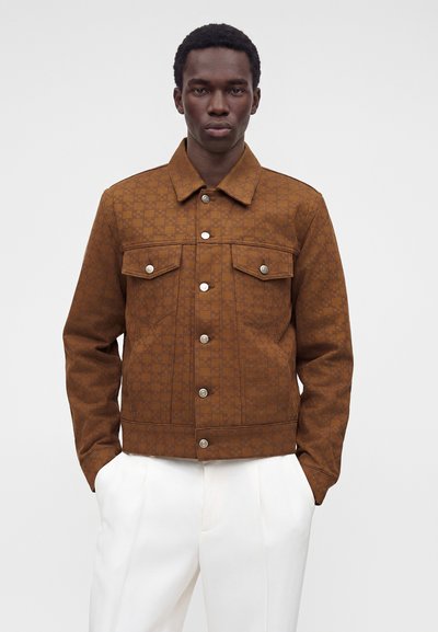 Homme portant une veste marron à carreaux boutonnée avec deux poches poitrine et un pantalon blanc, debout les mains dans les poches devant un fond uni.