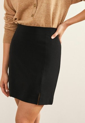Femme portant une jupe noire longueur genou avec une petite fente devant et un haut beige à manches longues avec des boutons au niveau de l'encolure.