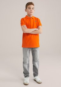 Pomarańczowa koszulka polo z kołnierzykiem i logo, w komplecie z jasnoszarymi dżinsami oraz białymi trampkami z szarymi i brązowymi akcentami. Stoi z założonymi rękami.