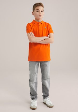 Oranžinė polo marškinė su apykakle ir logotipu, derinama su šviesiai pilkomis džinsomis ir baltais sportbačiais su pilkais ir rudais akcentais. Stovi su sukryžiuotomis rankomis.