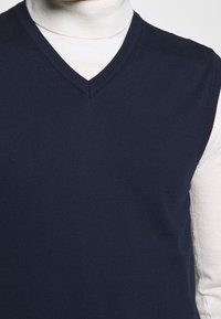 Marinblå stickad väst med V-formad halsringning, med ett texturerat tyg, lagd över en ljusfärgad polotröja. Enkelt, klassiskt snitt.