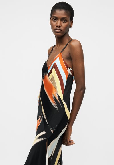 Γυναίκα που φοράει αμάνικο φόρεμα με λεπτές τιράντες και αφηρημένο μοτίβο σε μαύρο, πορτοκαλί, μπλε, κίτρινο και καφέ, πόζαρε μπροστά σε απλό φόντο.