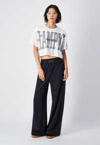 Gekürztes weißes Baumwoll-T-Shirt mit fettem grauen Text, kombiniert mit lockeren schwarzen Sweatpants, die ein Logo-Detail an der Seite aufweisen.